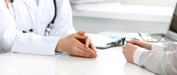 Close up of a doctors hands on a desk oposite those of a patient during conversation.