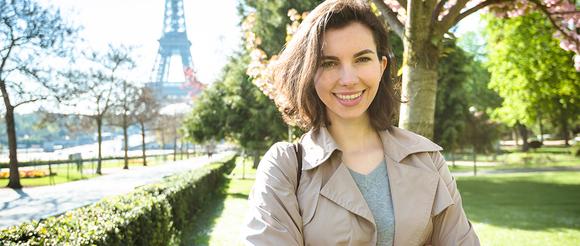 Female student in Paris.  She is standing in a park with the Eifel tower in the distance behind her.