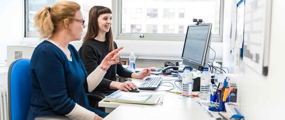 A student sitting at a desk using a pc.  Next to her is a tutor who is pointing at the screen and giving advice.