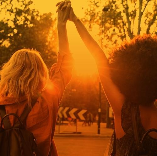 A sunset casts a golden hue as two women walk hand in hand down a street, symbolising friendship and connection.